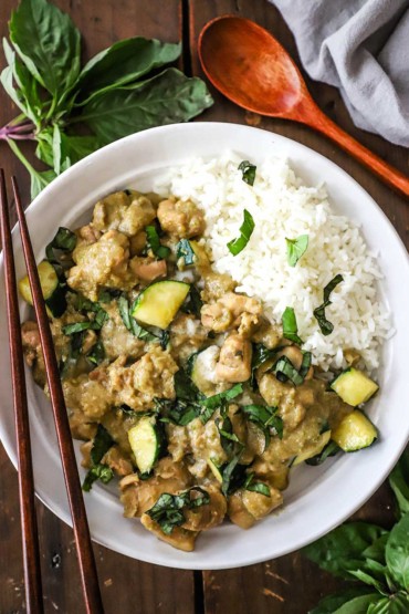 An overhead view of a shallow white bowl filled on one side with Thai green curry chicken and the other side with steamed Jasmine rice and topped with chopped Thai basil.