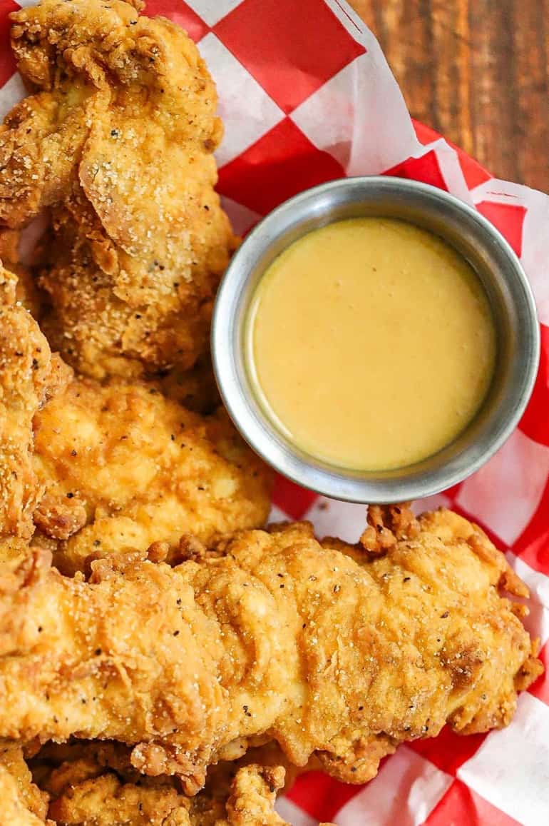 An overhead view of a silver condiment holder filled with homemade honey mustard dipping sauce surrounded by crispy chicken tenders.