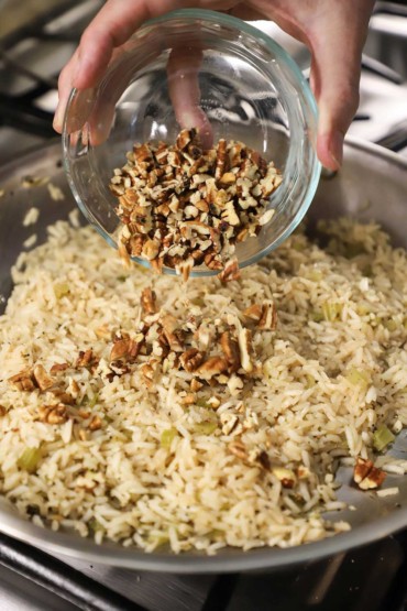 A person dumping roasted chopped pecans from a small glass bowl into a skillet of fully cooked rice pilaf.