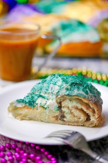 A close-up view of a slice of Mardi Gras King cake with glitter green icing on top and a circular cinnamon filling is visible.