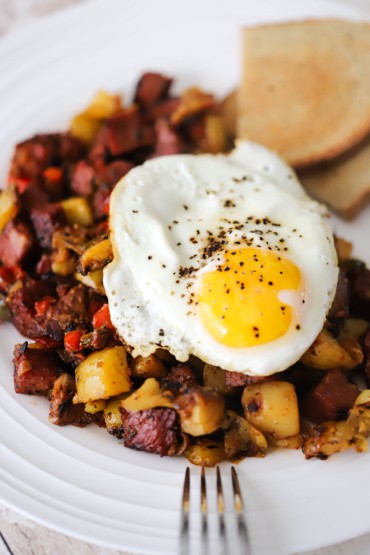 A close-up view of a fried egg sunny-side up sitting on a pile of corned beef hash all on a white plate with sliced rye bread in the background.