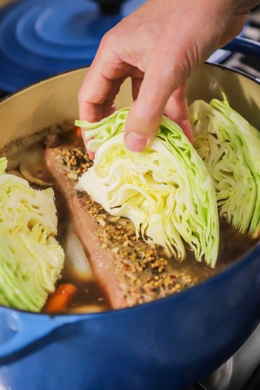 A person placing wedges of freshly cut green cabbage into a large blue Dutch oven that is filled with corned beef that is being braised in beef broth.