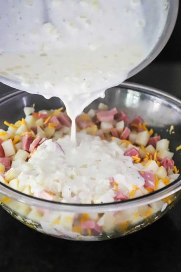 A creamy béchamel sauce being poured from a skillet into a large glass bowl filled with cubed potatoes, shredded cheddar cheese, and chunks of ham.