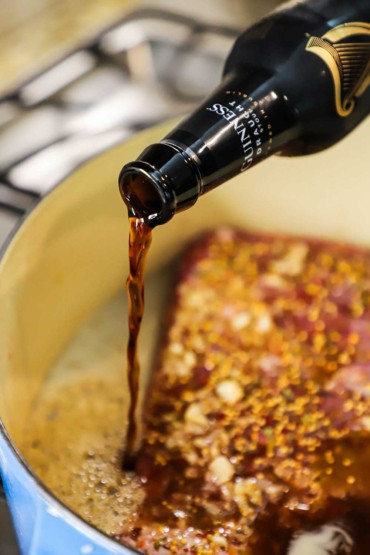 A person pouring a stout Guinness beer into a blue Dutch oven that has beef broth and a corned beef brisket that has been seasoned with mustard seeds and herbs.