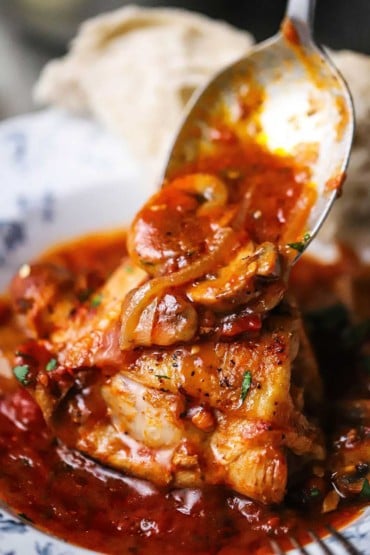 A large silver spoon being used to transfer a tomato, onion, and mushroom sauce onto seared chicken in a bowl.