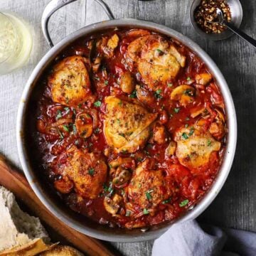 An overhead view of a large silver skillet filled with chicken cacciatore consisting of six seared chicken thighs in a thick tomato sauce.