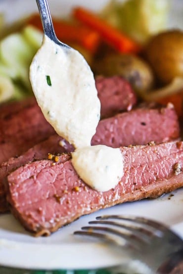A person using a spoon to drizzle a horseradish chive sauce over strips of braised corn beef on a white dinner plate.
