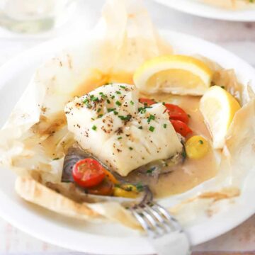 A straight-on view of two dinner plates that are holding servings of fish en papillote with a lemon wedge off to the side.
