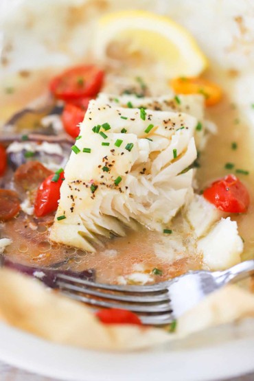 A close-up view of a fish en papillote that has been partially eaten and is resting in parchment paper with cooking liquid and steamed cherry tomatoes.