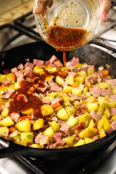 A person pouring a brown sauce with dried herbs into a cast-iron skillet that is filled with simmering cubed potatoes, corned beef, and vegetables.