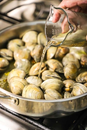 A person pouring white wine from a glass measuring cup into a skillet filled with unopened littleneck clams.