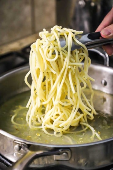 A person using a pair of tongs to place cooked spaghetti into a skillet filled with clam broth and sautéed garlic.