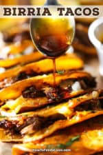 A straight on view of a person using a metal spoon to drizzle consommé over a row of beef birria tacos that are arranged side by side on a board.
