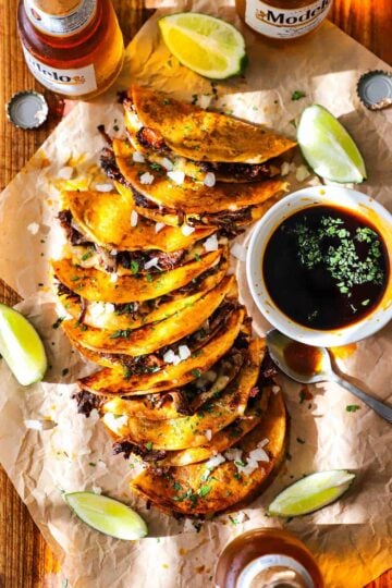 An overhead view of a row of beef birria tacos that are on laying on their sides next to a white dipping bowl filled with beef consommé and surrounded by lime wedges and Mexican bottled beer.
