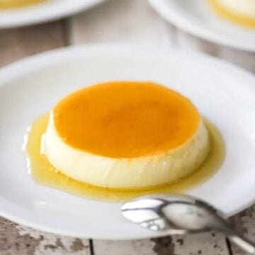 A straight-on view of homemade flan that has a light caramel sauce on top of it and surrounding the base and is on a white plate with other plated servings nearby.