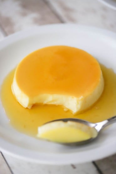 A front-view of a homemade flan that is on a white dessert plate with a light caramel sauce on top of and surrounding the partially eaten dessert.