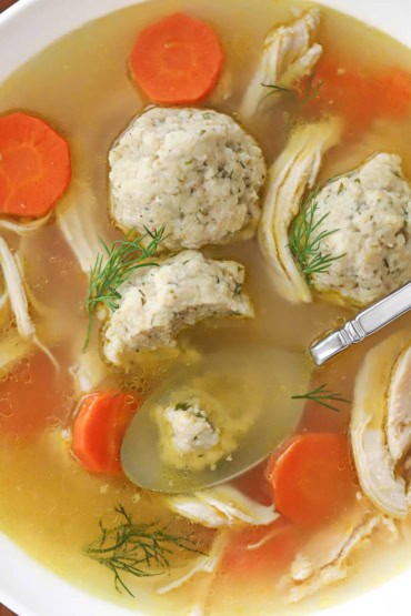An overhead view of a bowl of matzo ball soup with a spoon in the soup with a partially eaten matzo ball on it.