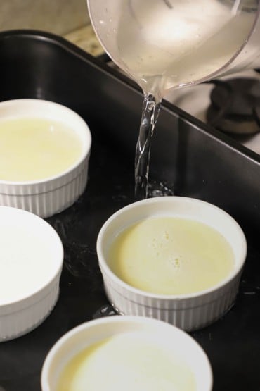A person pouring hot water from a large pitcher into a black roasting pan that has four ramekins in it that are partially filled with vanilla custard.
