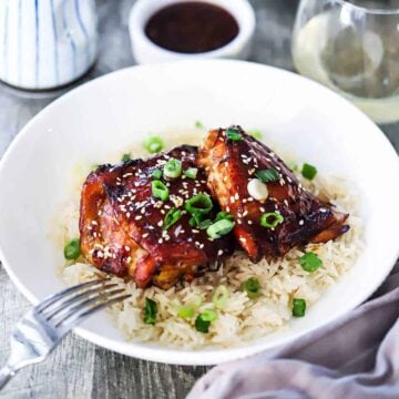 A close-up view of a two pieces of baked teriyaki chicken thighs resting on a bed of Jasmine rice and garnished with sesame seeds and chopped green onions.