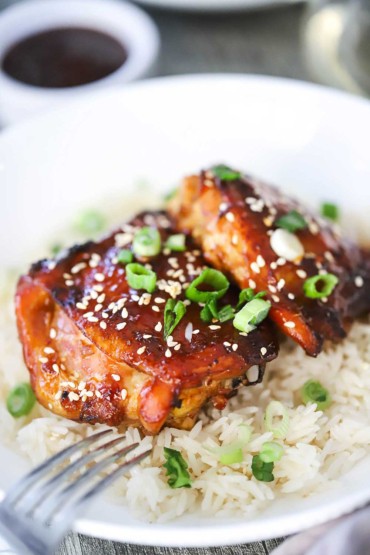 A close-up view of a two pieces of baked teriyaki chicken thighs resting on a bed of Jasmine rice and garnished with sesame seeds and chopped green onions.
