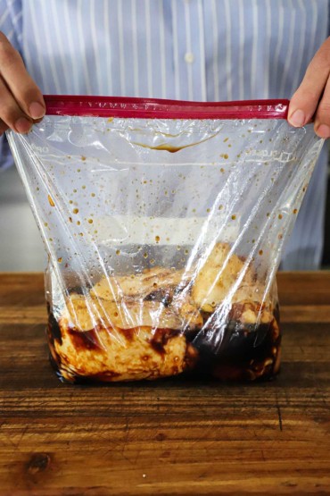 A person holding a large plastic baggie that is filled with uncooked skin-on chicken thighs that are soaking in teriyaki marinade.