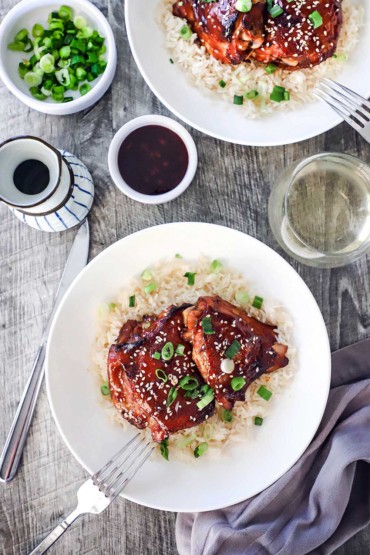 An overhead view of a white bowl filled with a serving of baked teriyaki chicken thighs sitting next to a glass of white wine and a small bowl of teriyaki sauce.
