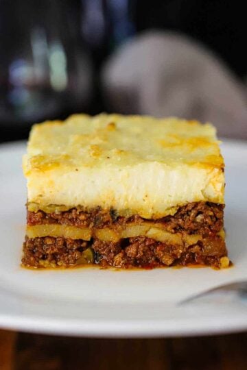 A straight-on view of a perfectly square piece of homemade moussaka sitting on a white dinner plate.