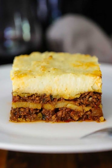 A straight-on view of a perfectly square piece of homemade moussaka sitting on a white dinner plate.