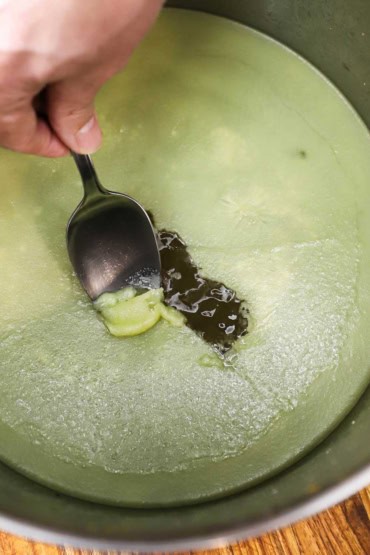 A person using a spoon to scrape congealed chicken fat, also known as schmaltz, from the surface of chilled chicken broth in a stock pan.