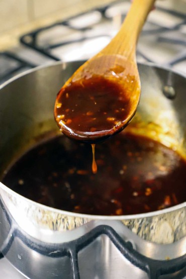 A large wooden spoon being used to raise a spoonful of teriyaki sauce from a medium-sized silver saucepan.