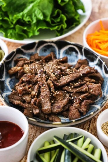 A close-up view of a shallow blue bowl that is filled with cooked bulgogi beef strips and surrounded by other small bowls of pickled vegetables and lettuce leaves.