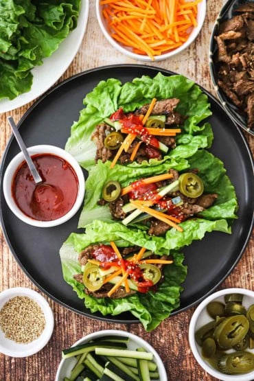 An overhead view of three beef bulgogi lettuce wraps that are sitting on a black plate and next to a small bowl filled with Korean barbecue sauce.