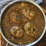 An overhead view of a large silver saucepan filled with four salisbury steaks that are resting in a mushroom brown gravy with a wooden spoon resting nearby.