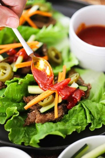 A person using a small spoon to drizzle Korean barbecue sauce over strips of bulgogi beef nestled into a lettuce leaf and toped with julienned carrots and cucumber.