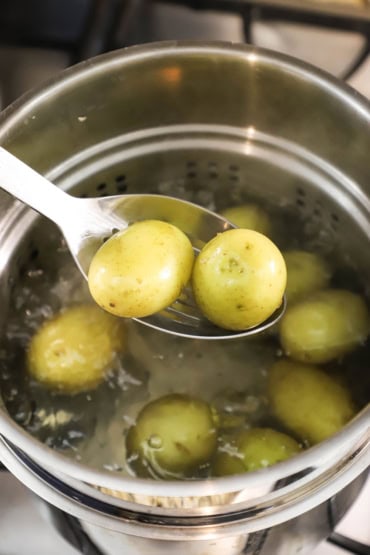 A person holding up a metal slotted spoon holding two medium-sized gold potatoe that have been removed from a pot of boiling water with other potatoes in it.