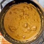 An overhead view of a black pot filled with chicken korma with a orange checkered napkin nearby.