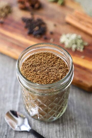 A close-up view of a small glass jar filled with freshly ground garam masala next to a cutting board topped with various spices.