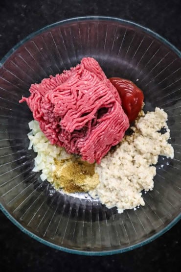 An overhead view of a glass bowl filled with milk-soaked breadcrumbs, ground beef, sautéed onions, ketchup, and seasonings.