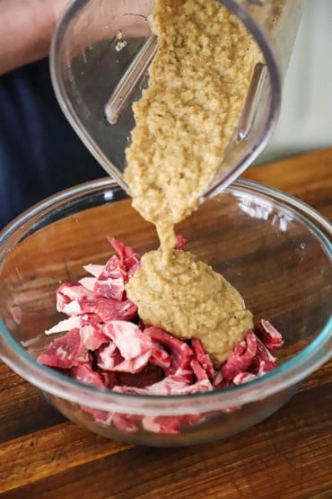 A person pouring a puréed pear and onion marinade from a blender into a glass bowl that is filled with thinly sliced pieces of beef.