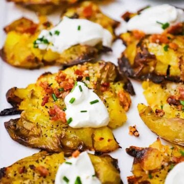 A close-up view of cooked smashed potatoes that are brown and crispy on the edges that have been topped with crumbled bacon, a dollop of sour cream, and snipped chives.