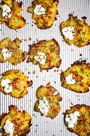An overhead view of a baking sheet filled with baked smashed potatoes that have been garnished with crumbled bacon, sour cream, and snipped chives.