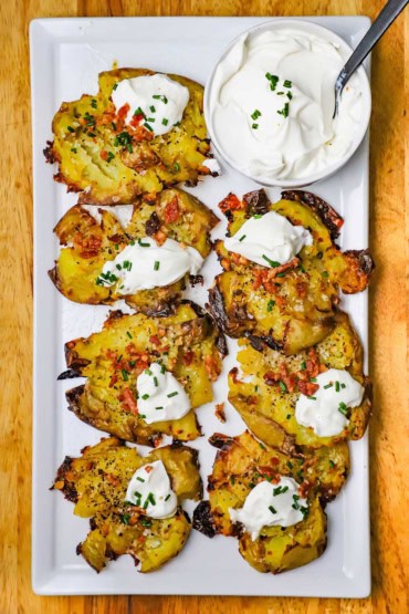 An overhead view of crispy smashed potatoes arranged on a white rectangular platter next to a small bowl filled with sour cream and topped with snipped chives.