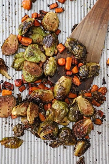 An overhead view of halved Brussels sprouts and pieces of carrots that have been tossed with balsamic vinegar and herbs and roasted on a large silver baking sheet.