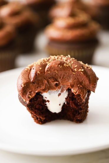 A half eaten chocolate cupcake with marshmallow filling sitting on a white dessert plate with the cream filling visible.