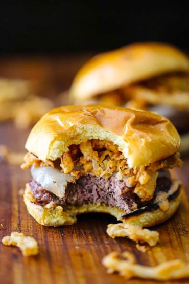 A straight-on view of a smoked steakhouse burger that is topped with fried onion strips that has a bite taken out of it and is sitting on a cutting board.