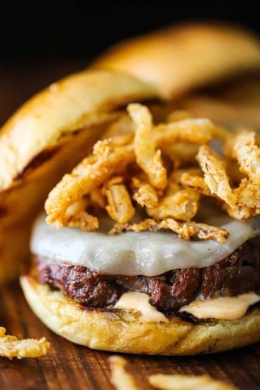 A close-up view of a smoked steakhouse burger that has a pile of fried onion strips sitting on top of melty provolone cheese and the top bun resting off to the side.