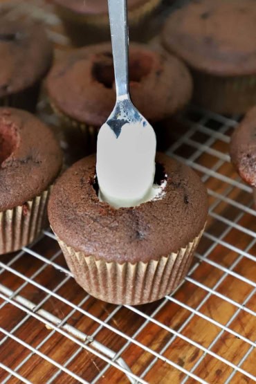A person using a small spoon to transfer marshmallow cream into the chocolate cupcake that has had a small well removed from its top.