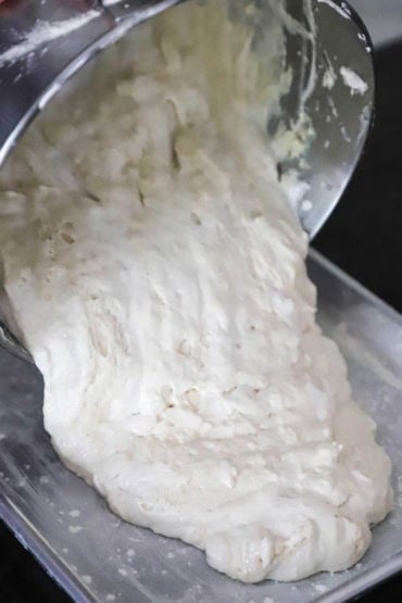 A person transferring a dense white cake batter from a mixing bowl into a cake pan that has been greased and dusted with flour.