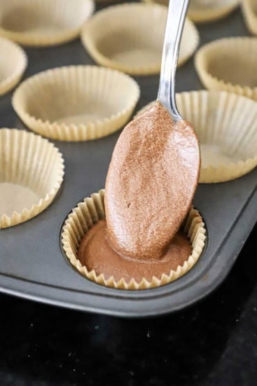 A person using a large spoon to transfer chocolate cupcake batter into a muffin tin that is lined with paper cupcake liners.