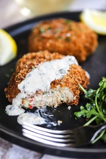 A close-up view of a half-eaten New England-style crab cake that has a strip of homemade tartar sauce on it all on a black dinner plate.